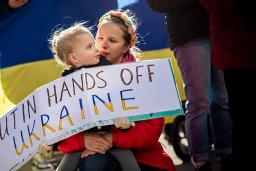 A child is holding a protest sign that says: “Putin, hands off Ukraine.” The child is sitting on their caregiver’s lap. Behind them is a Ukrainian flag. 