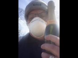 A self-shot photo of a man wearing a toque and a disposable breathing mask holding a bottle of sparkling wine.