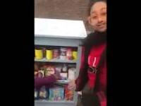 A woman stands outside next to a cabinet holding a variety of canned and packaged food.