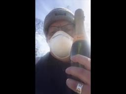A self-shot photo of a man wearing a toque and a disposable breathing mask holding a bottle of sparkling wine.