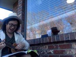 A man wearing a hat playing a ukulele on a front porch, outside a large picture window.
