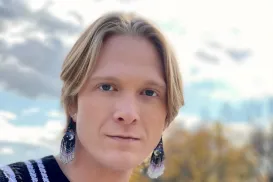 A blond male man wearing beaded earrings and a black- and- white vertically striped shirt.