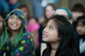 A girl lifts her head and eyes slightly upward. She has a smile on her face. Behind her are more children smiling.