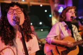 Two women wearing glasses and white shirts are singing and playing their guitars.