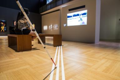 In the foreground, a visually impaired person’s white stick touches three tactile strips laid out on the floor of a Museum gallery. The strips lead to benches, where two people are watching a film on a large screen.