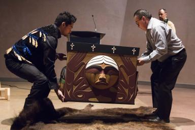 Luke Marston (left) and another person lift the Bentwood Box from a buffalo hide, which lies on the floor underneath.