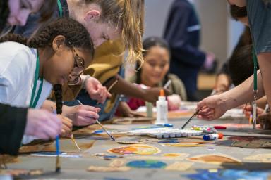 A group of children and youth paint with small brushes. They are gathered on both sides of a long table with different coloured paints in the foreground.