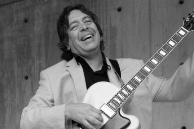 A black- and- white shot of a male musician standing up, laughing and tuning his guitar.