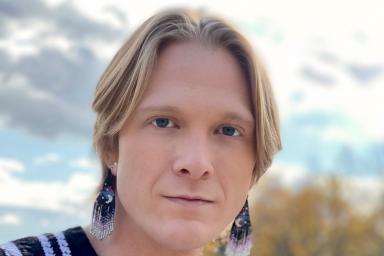 A blond male man wearing beaded earrings and a black- and- white vertically striped shirt.