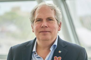 Un homme se tient face à l’objectif avec une expression sérieuse. Il porte une veste de costume sombre et une chemise boutonnée bleu clair. Il arbore une fleur multicolore de style métis sur son revers.