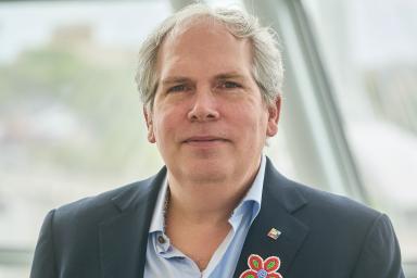 A man stands facing the viewer with a serious expression. He is wearing a dark suit jacket and a light blue buttoned-up shirt. He sports a multicoloured Métis-style flower on his lapel.