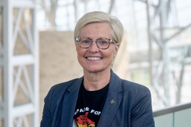 A woman with light hair and dark glasses smiles at the viewer. She is wearing a dark suit jacket and t-shirt that features a map of Canada in the colours of the inclusive Pride flag.