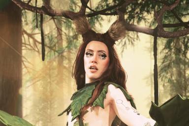 A drag artist is dressed in a cosplay costume with long, brown hair; large antlers; and green leaf scarf, skirt and gloves.
