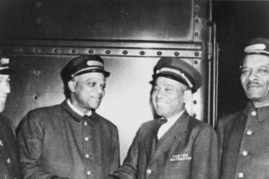 A black and white photo of four men in train porter uniforms. All of the men are smiling, and the two men in the middle appear to be shaking hands.