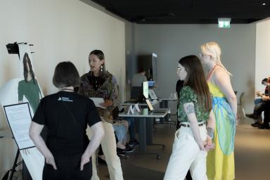 In a museum gallery, a student talks to three people next to a life-size drawing of themself.