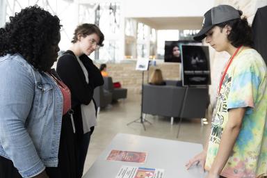 One student and two adults stand on opposite sides of a table with posters on it.