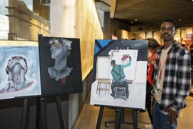 In a museum gallery, a student stands beside three paintings that they've created.