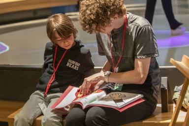 An older and a younger student sitting beside each other on a bench. The older student is reading a book to the younger student. 