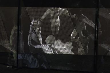 A still from black-and-white film, seen through a heavy black net, depicts dancers dressed in military uniforms forming a circle, holding hands.