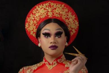 A drag artist is wearing Vietnamese cultural clothing with a matching red and gold headdress and long dress.