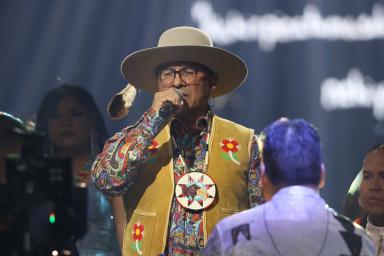 Close-up on a group of people wearing beaded jackets and vests performing on stage. In the centreer, a man wearing glasses, a hat, and a large, beaded medallion sings into a microphone.