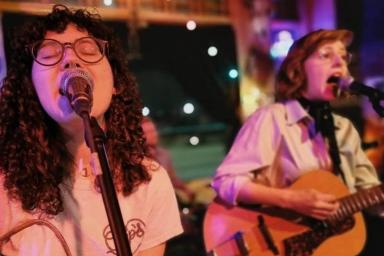 Two women wearing glasses and white shirts are singing and playing their guitars.
