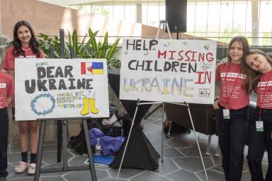 On a sunny day, four students stand on either side of two easels. The students wear matching red shirts that say “Be an Upstander” and the signs on the easels say “Dear Ukraine” and “Help Missing Children in Ukraine.”