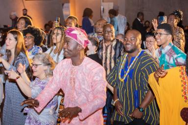 A group of people of all ages, many in brightly coloured traditional African attire, dancing and smiling.