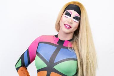 A drag artist with long blond hair is dressed in a colourful one-piece accented with thick black lines.