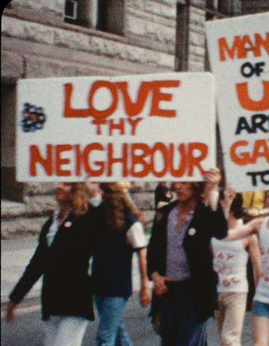 A group of people march down a city street. At the front of the group, on the left side of the image, two people hold a sign over their heads that reads “Love thy neighbour.” Slightly behind them is a sign that reads “Many of u are gay too!” Partially obscured.