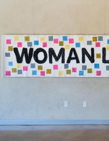 A white banner hangs on a wall, bearing the words "Woman, Life, Freedom" in black. Small coloured squares with text are attached to the banner around the text. Partially obscured.