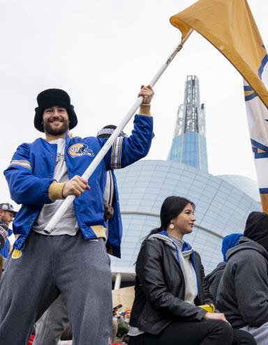 A smiling man in a crowd of people holding a large yellow flag with a blue and white, “W” in the centre. The man is wearing a blue Winnipeg Blue Bombers jacket as he stands in front of the Canadian Museum for Human Rights. Partially obscured.