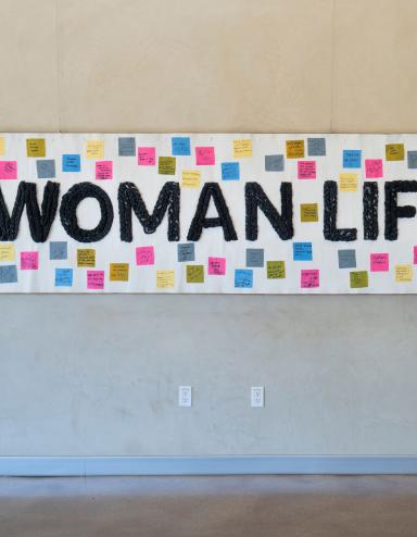 A white banner hangs on a wall, bearing the words "Woman, Life, Freedom" in black. Small coloured squares with text are attached to the banner around the text. Partially obscured.