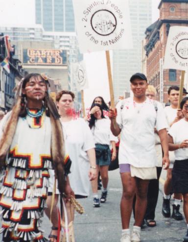 A group of marchers carrying signs that read “2-Spirited People of the 1st Nations” are walking down the centre of a city street while onlookers watch behind barriers on both sides. Partially obscured.