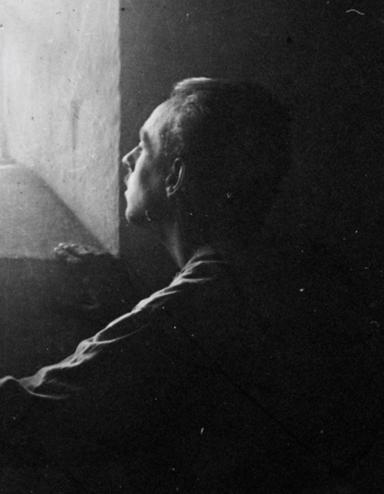 A man stands in dark shadows in a stark stone cell, gazing upward at bright sunlight streaming in through a barred window. Partially obscured.