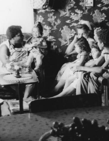 A black and white image of women and young children sitting on couches and chairs in a small living room. Partially obscured.