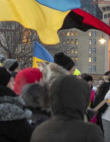 Une foule de personnes emmitouflées se tient debout dans une rue de ville pendant l’hiver, tenant de multiples drapeaux ukrainiens bleus et jaunes ainsi qu’un drapeau canadien. Le rassemblement semble être une manifestation en appui à l’Ukraine. De grands édifices à bureaux et des feux de circulation sont à l’arrière-plan sous un ciel couvert. Partially obscured.