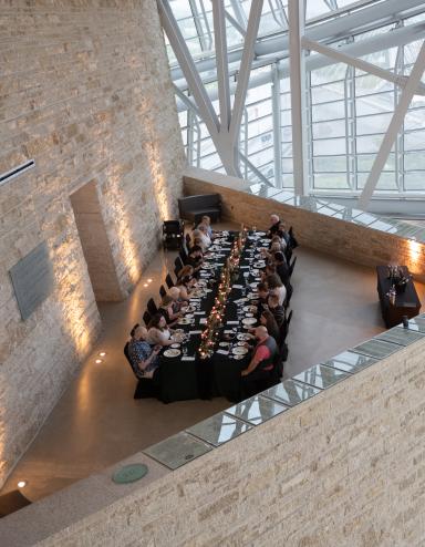 An elegant long table with people dining in an architecturally striking space, featuring tall glass windows and a high ceiling supported by metal beams. Partially obscured.