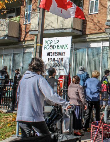 A line-up of people wait outside a building. There are signs indicating it is a Food Bank. A Canadian flag flies above them. Partially obscured.