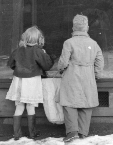 Black-and-white photograph of two children looking into an abandoned store window. Partially obscured.