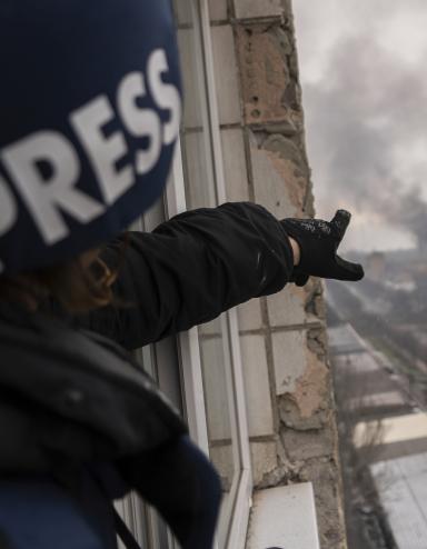 A person wearing a dark blue helmet marked with the word “PRESS” points from a balcony window high above a road, towards a pillar of smoke following an explosion. Partially obscured.