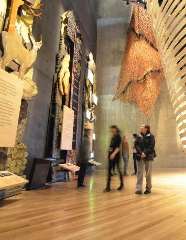 Five museum visitors look at towering exhibits in the “Indigenous Perspectives” gallery at the Canadian Museum for Human Rights. The nearest is made of wood and features trees, animals and a plaque. Behind the visitors, a rounded theatre built of bent wooden slats is visible. Partially obscured.