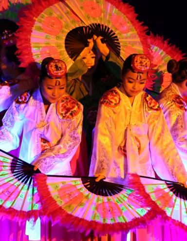 Korean dancers holding large, flower-patterned fans are grouped together so their fans form a circle. Partially obscured.