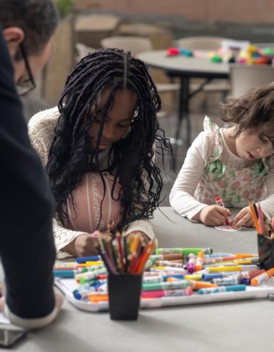 Two children sit at a table making art while an adult leans on the table, watching. Partially obscured.