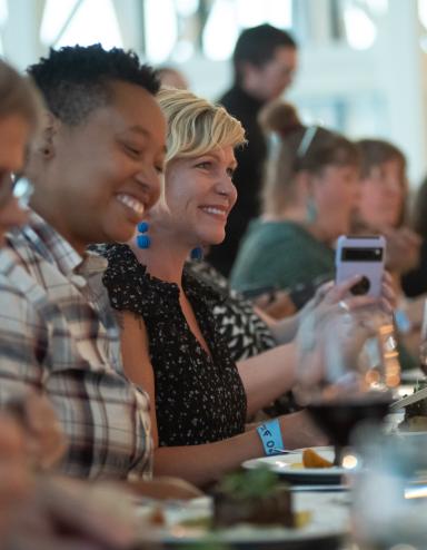 A group of people sit at a long dining table, laughing and enjoying a meal together. The focus is on two people smiling brightly - one wearing a plaid shirt and the other in a black patterned top, with glasses and plates arranged neatly in front of them. Partially obscured.
