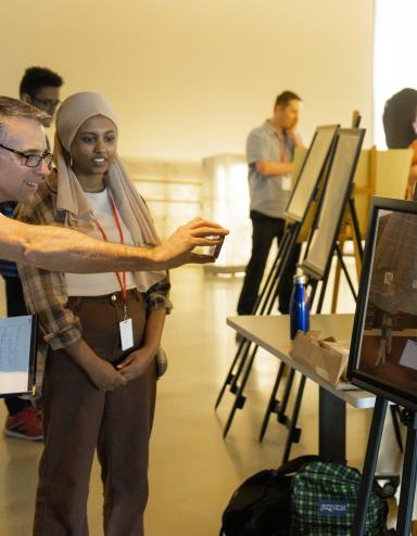Students in a museum gallery display their work on easels. The focus is on one student talking with an adult who is smiling and taking a photograph with their phone of the student’s work. Partially obscured.