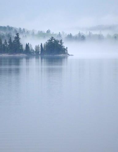 Mist rises off a large, calm lake surrounded by an evergreen forest. Partially obscured.