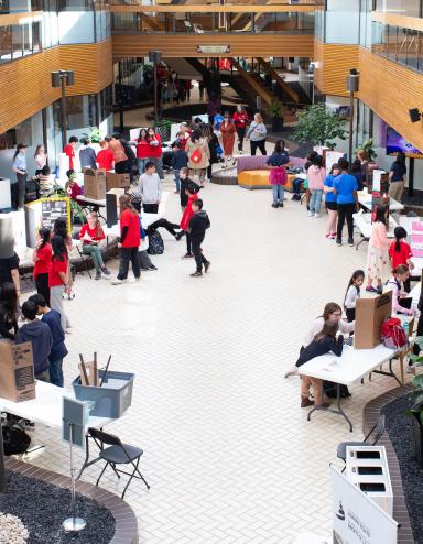Many people gathered in an open indoor space with tables and posters set up on them. Partially obscured.