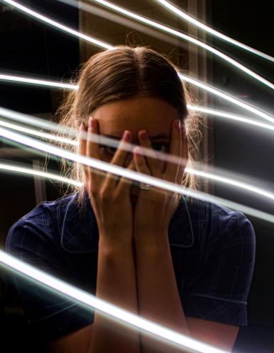 A teen girl holds her hands over her face and looks out stricken through her fingers. Bright while lines swirl around her. Partially obscured.