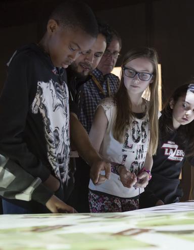 A Museum guide showing five students how to engage with the digital table, around which they are all standing. Partially obscured.